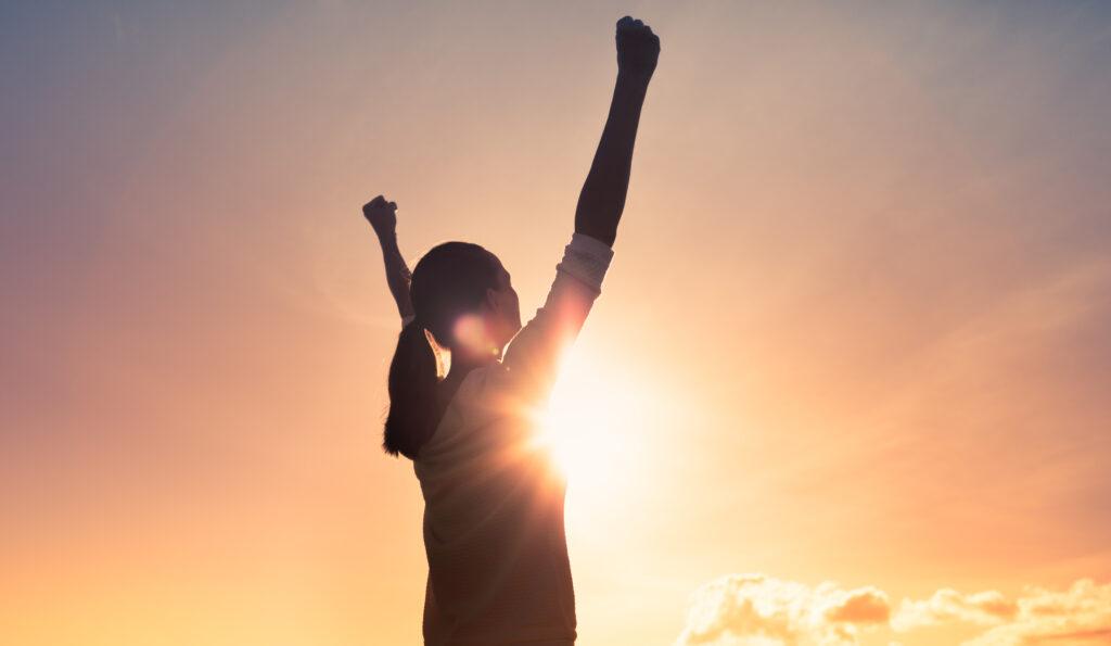 Woman raising arms in celebration with sunrise in background.
